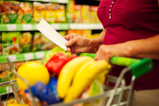 A person holding a shopping list in one hand while pushing a supermarket trolley filled with bananas, peppers, and other fresh groceries.