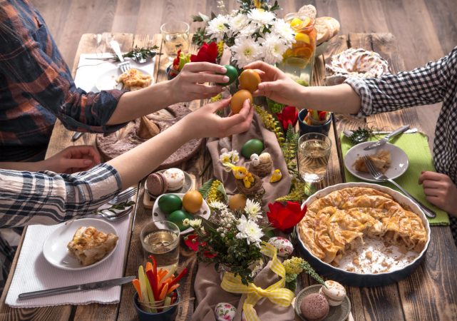 Several people gathered around a rustic table decorated with flowers, colored eggs, pastries, and a pie, reaching in to tap dyed Easter eggs together during a festive meal.